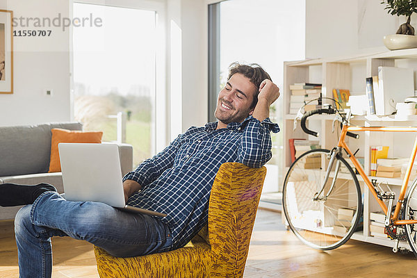 Porträt des lächelnden jungen Mannes auf Sessel im Wohnzimmer mit Laptop