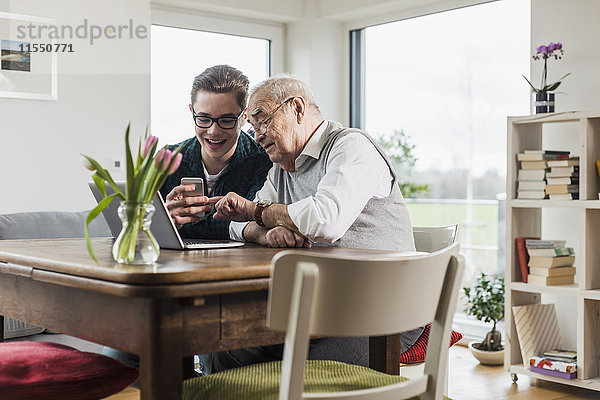 Senior Mann und sein Enkel sitzen am Tisch im Wohnzimmer mit Laptop und Smartphone