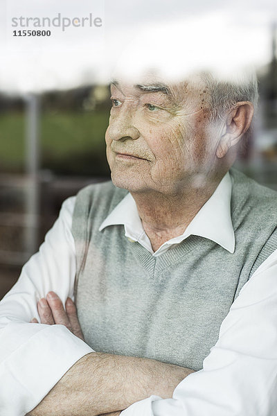 Porträt eines nachdenklichen älteren Mannes mit gekreuzten Armen  der durchs Fenster schaut.