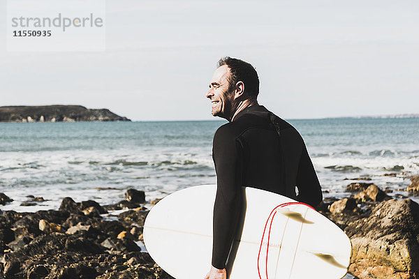 Frankreich  Bretagne  Finistere  Halbinsel Crozon  lächelnder Mann am Felsenstrand mit Surfbrett