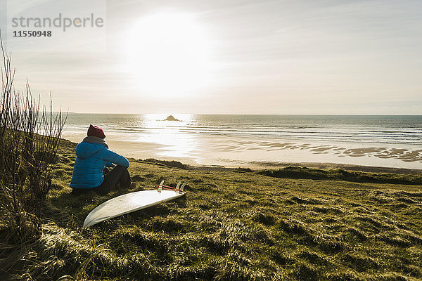 Frankreich  Bretagne  Finistere  Halbinsel Crozon  Frau bei Sonnenuntergang an der Küste sitzend mit Surfbrett