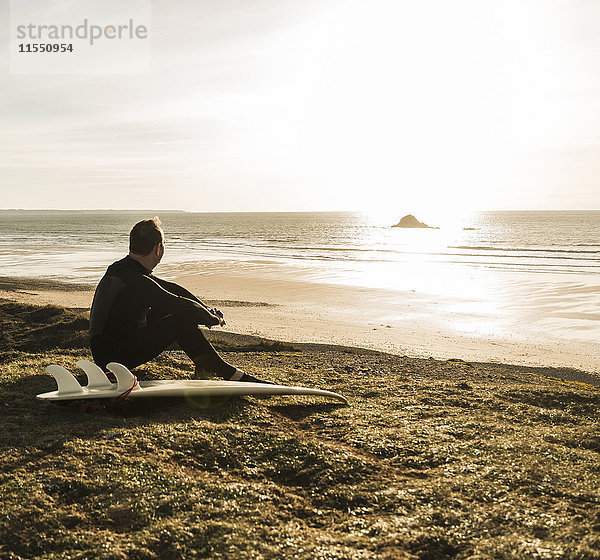 Frankreich  Bretagne  Finistere  Halbinsel Crozon  Mann bei Sonnenuntergang an der Küste sitzend mit Surfbrett