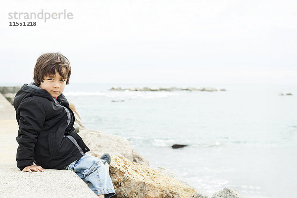 Portrait eines kleinen Jungen  der auf einer Wand vor dem Meer sitzt.