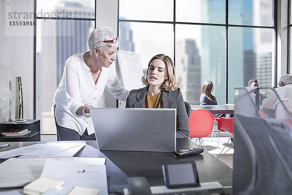 Zwei Frauen im Büro mit Laptop und Besprechung im Hintergrund