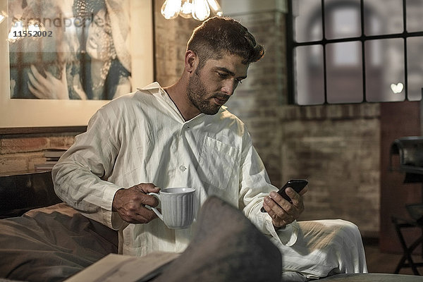 Mann sitzt auf dem Bett und schaut auf das Telefon und trinkt Kaffee.
