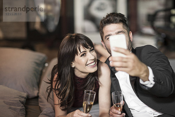 Lächelndes Paar in eleganter Kleidung trinkt Champagner im Bett und nimmt einen Selfie.