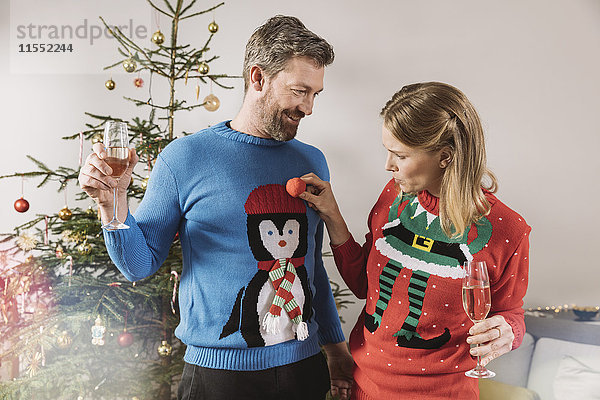 Zwei Leute mit hässlichen Weihnachtspullovern  die vor dem Baum lachen.