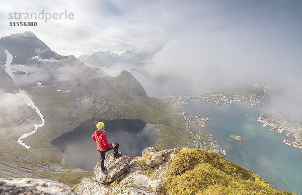 Wanderer auf dem Gipfel bewundert den blauen See und das Meer  die das Dorf umrahmen  Reinebringen  Moskenesoya  Lofoten  Norwegen  Skandinavien  Europa