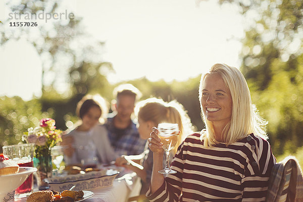 Lächelnde Frau trinkt Wein am sonnigen Gartenpartytisch