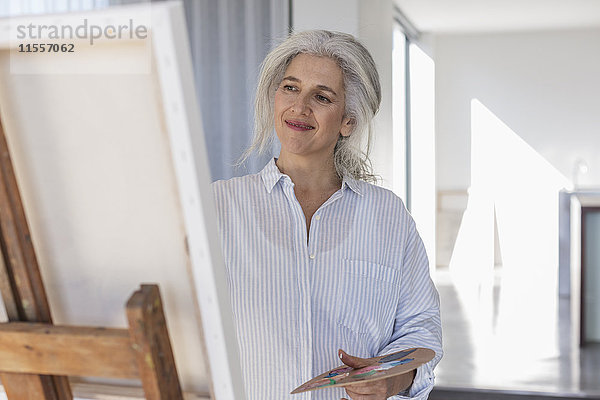 Lächelnde reife Frau mit Palette malt an Leinwand auf Staffelei