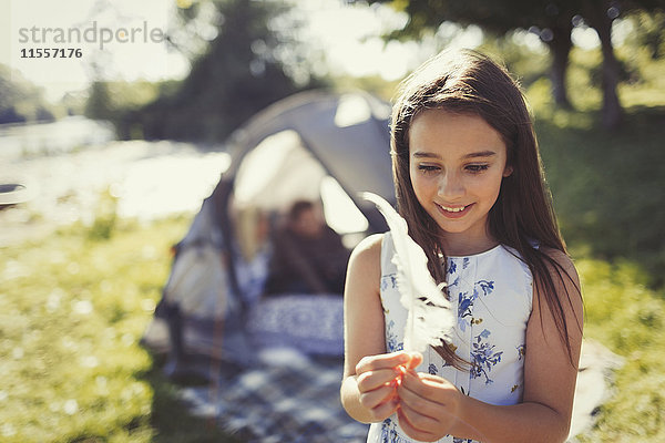 Lächelndes Mädchen hält weiße Feder vor dem sonnigen Zelt auf dem Campingplatz