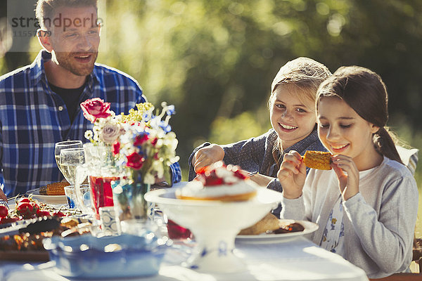 Vater beobachtet Töchter beim Essen am sonnigen Gartenparty-Tisch