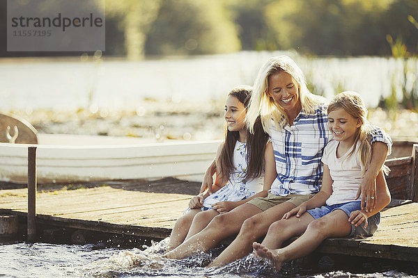 Lächelnde  liebevolle Mutter und Töchter auf dem Dock und spritzende Füße im See.