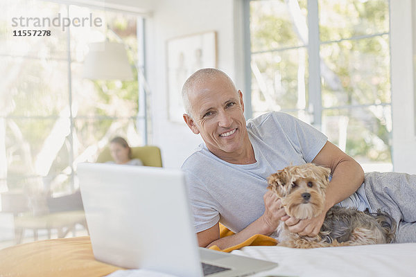 Porträt lächelnder Mann  der einen Hund am Laptop auf dem Bett streichelt