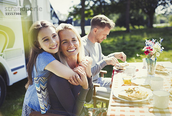 Lächelnde  liebevolle Mutter und Tochter  die sich am Tisch vor dem sonnigen Wohnmobil umarmen.