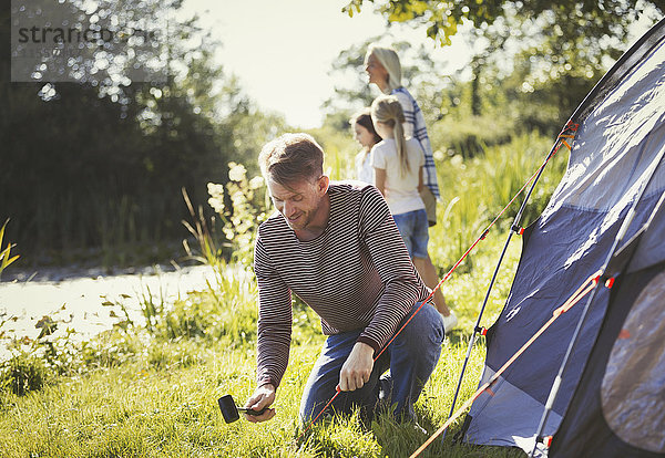 Vater schlägt Zelt auf einem sonnigen Campingplatz am See auf.