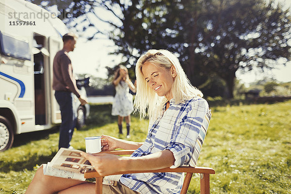 Frau liest Magazin und trinkt Kaffee draußen im sonnigen Wohnmobil