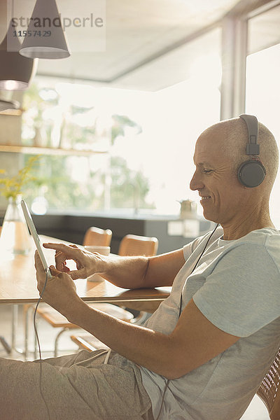 Älterer Mann  der mit Kopfhörern und digitalem Tablet am Esstisch Musik hört