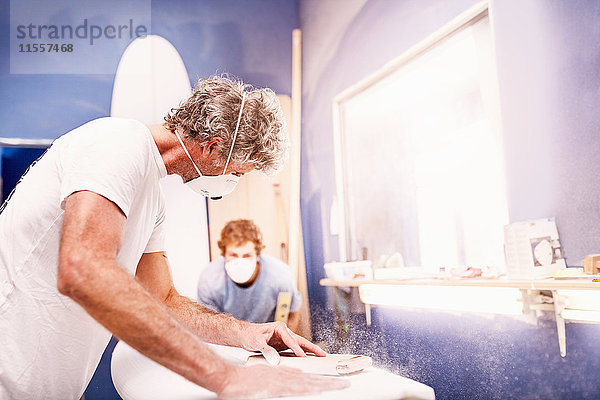 Männliche Surfboard-Designer mit Schutzmaske beim Schleifen von Surfbrettern in der Werkstatt