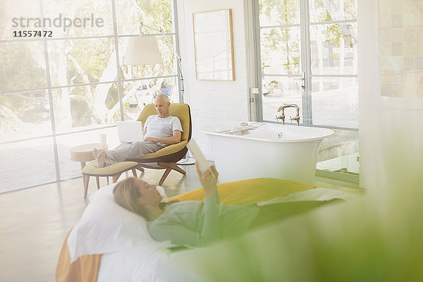 Älteres Paar entspannt sich mit Laptop und digitalem Tablet in einem luxuriösen Schlafzimmer