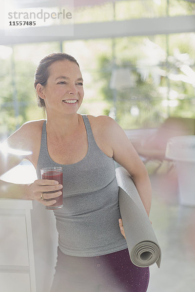 Lächelnde reife Frau mit Yogamatte trinkt gesunden Frucht-Smoothie
