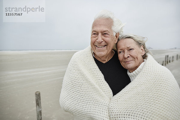 Lächelndes  liebevolles Seniorenpaar in eine Decke am Strand gewickelt