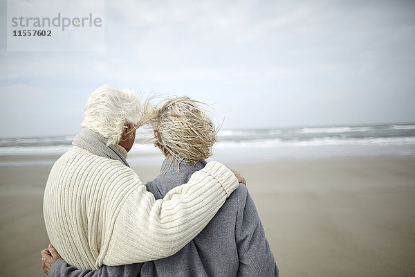 Nachdenkliches Seniorenpaar beim Umarmen und Blick aufs Meer am windigen Winterstrand