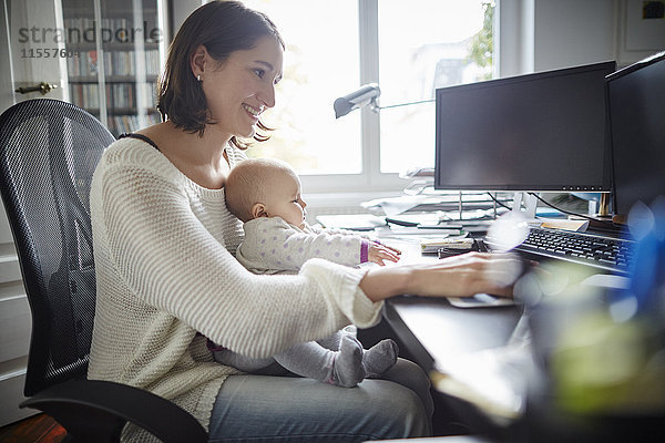 Lächelnde Mutter mit Baby-Tochter am Schreibtisch im Home-Office
