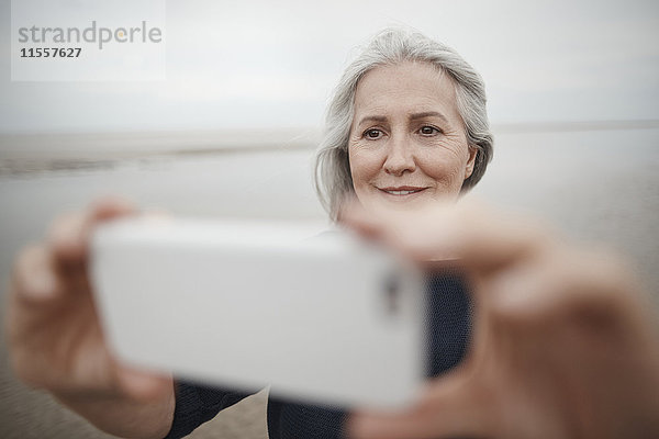 Seniorin nimmt Selfie mit Fotohandy am Winterstrand mit.