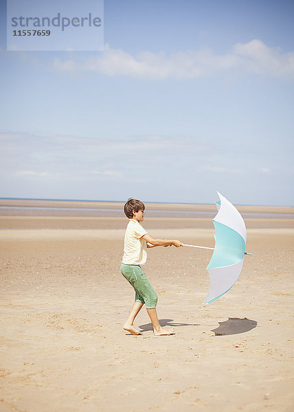 Wind zieht Regenschirm in den Händen eines Jungen am sonnigen Sommerstrand