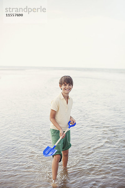 Portrait lächelnder Junge mit Schaufel in der Meeresbrandung am Sommerstrand