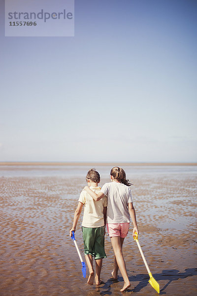 Bruder und Schwester mit Schaufeln  die sich umarmen und im nassen Sand am sonnigen Sommerstrand unter blauem Himmel laufen