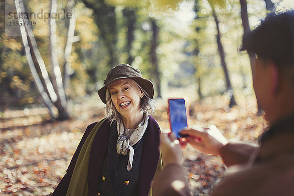 Verspielte Seniorin beim Fotografieren durch den Ehemann mit Fotohandy im Herbstpark