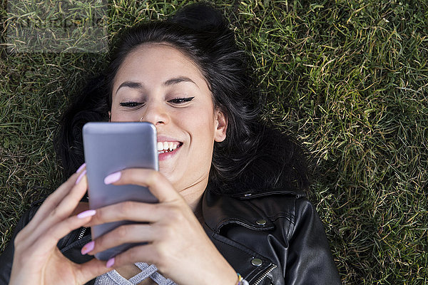 Porträt einer lächelnden jungen Frau mit Nasenpiercing auf der Wiese liegend mit Blick auf das Handy