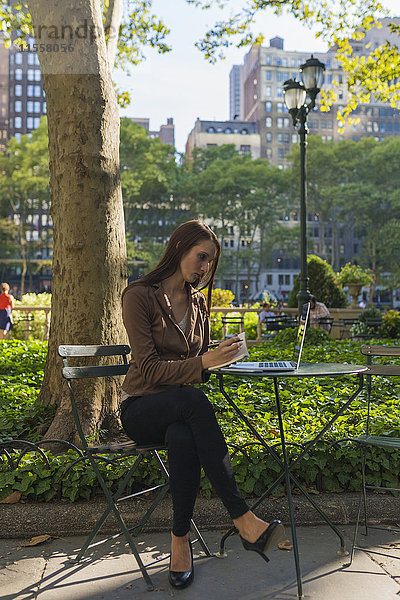 USA  New York  Manhattan  junge Geschäftsfrau mit Laptop am Tisch im Park und Notizen machend