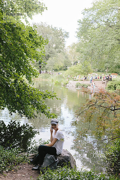 Junge Frau sitzt am Seeufer im Park und telefoniert mit dem Handy.