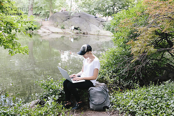 Junge Frau sitzt am Seeufer im Park mit Laptop