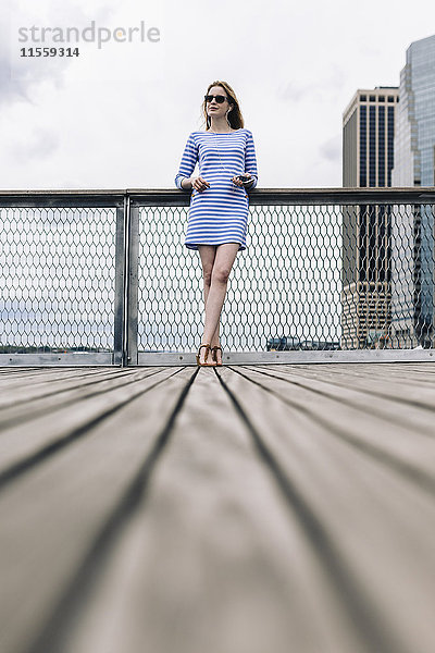 USA  New York City  Junge Frau steht in Manhattan am Geländer und hält ein Smartphone.