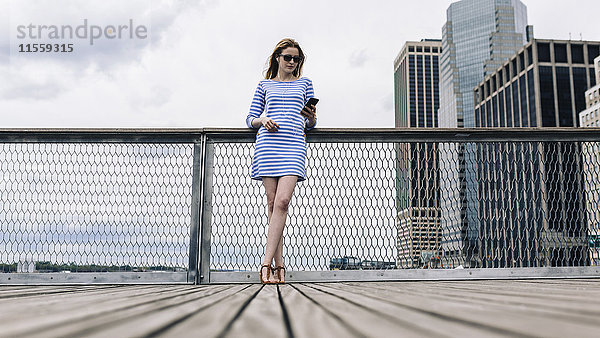 USA  New York City  Junge Frau steht in Manhattan am Geländer und hält ein Smartphone.