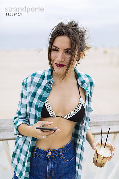 Junge Frau am Strand mit Tasse Kaffee und Smartphone