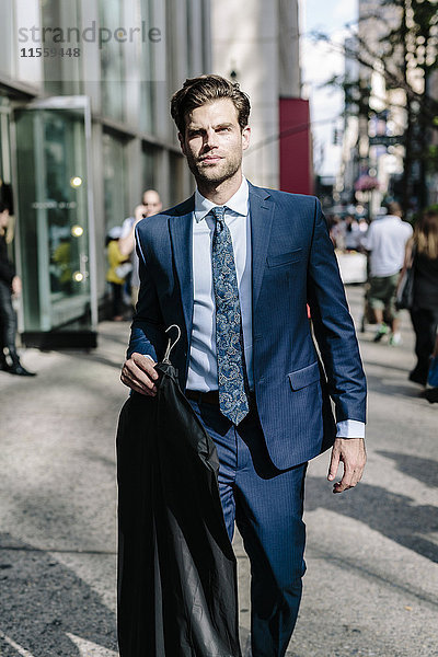 Ein gutaussehender Geschäftsmann  der in Manhattan spazieren geht und eine Jacke auf einem Kleiderbügel trägt.