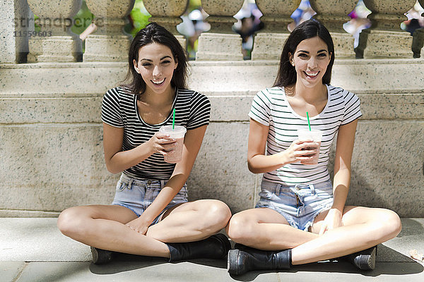 Zwei glückliche Zwillingsschwestern sitzen im Freien und trinken etwas zum Mitnehmen.