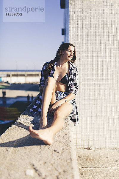 Junge hübsche Frau in Strandkleidung an der Wand sitzend