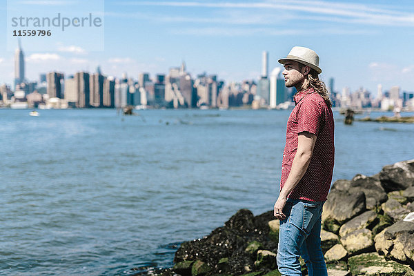 USA  New York City  Mann mit Hut am Wasser mit Skyline von Manhattan im Hintergrund