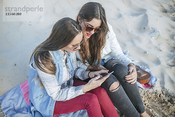 Zwei junge Frauen sitzen am Strand und schauen auf das Handy.