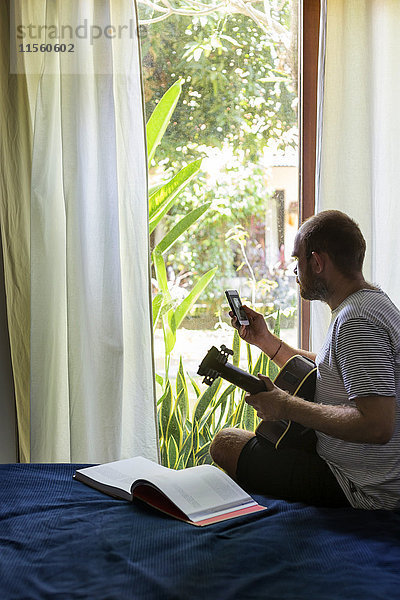 Mann mit Gitarre und Handy auf dem Bett sitzend