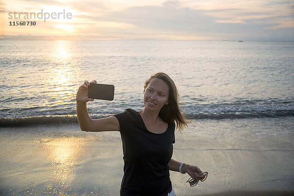 Indonesien  Bali  Frau mit einem Selfie am Strand bei Sonnenuntergang