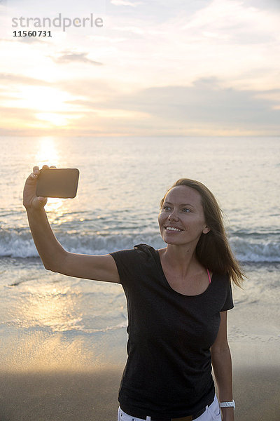 Indonesien  Bali  Frau mit einem Selfie am Strand bei Sonnenuntergang