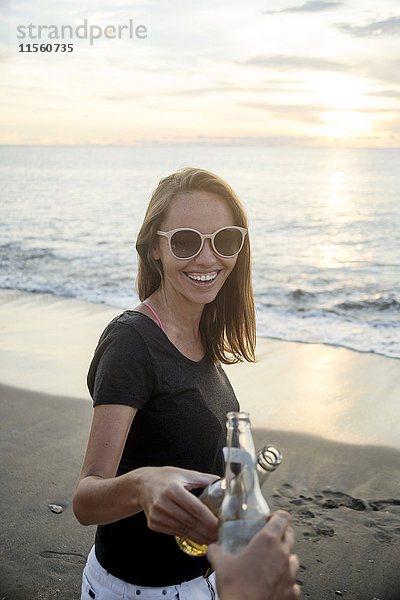 Indonesien  Bali  glückliche Frau klirrende Bierflasche mit Freund am Strand bei Sonnenuntergang