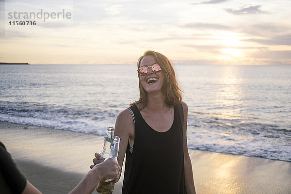 Indonesien  Bali  glückliche Frau klirrende Bierflasche mit Freund am Strand bei Sonnenuntergang